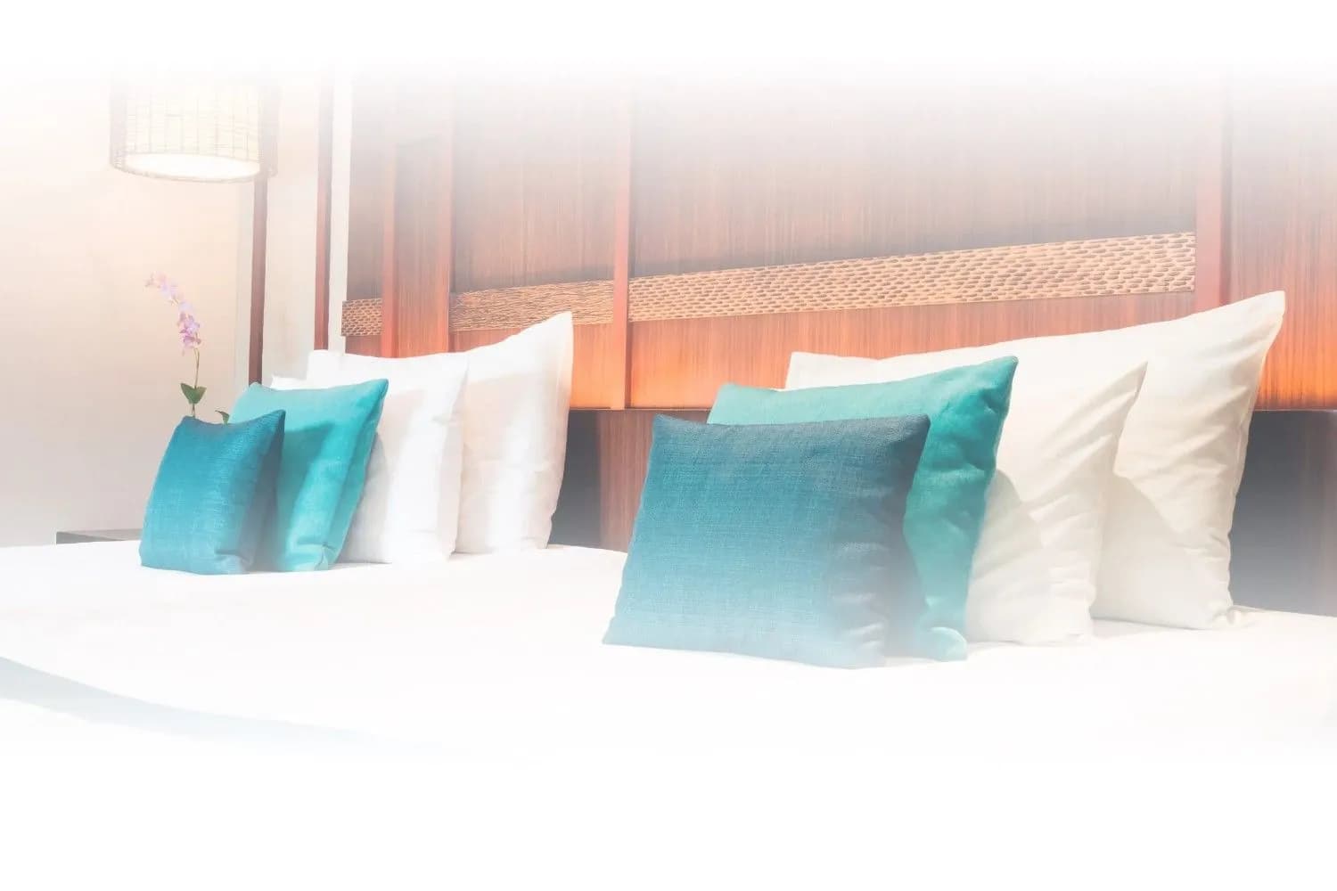 A made bed with crisp white linen and teal cushions against a warm timber headboard