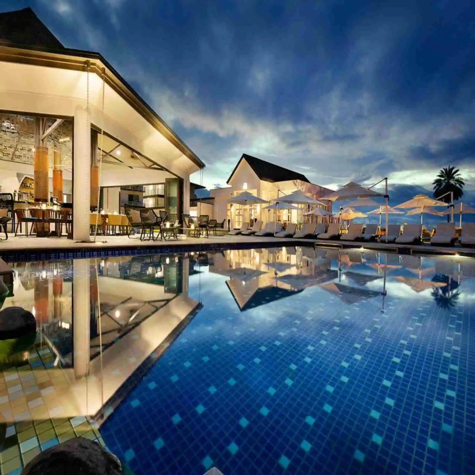 A hotel's open-sided restaurant and sun loungers lit up at dusk beside a long blue pool
