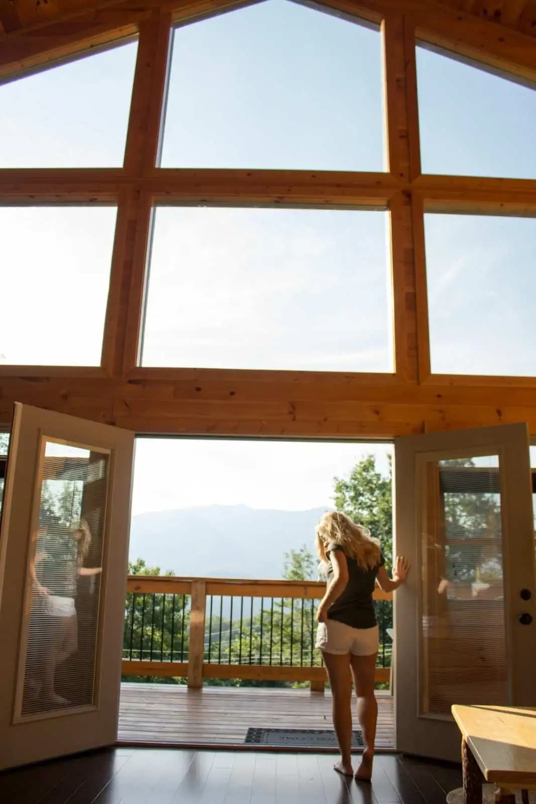 A guest opening the doors of a timber lodge onto a deck above forested hills